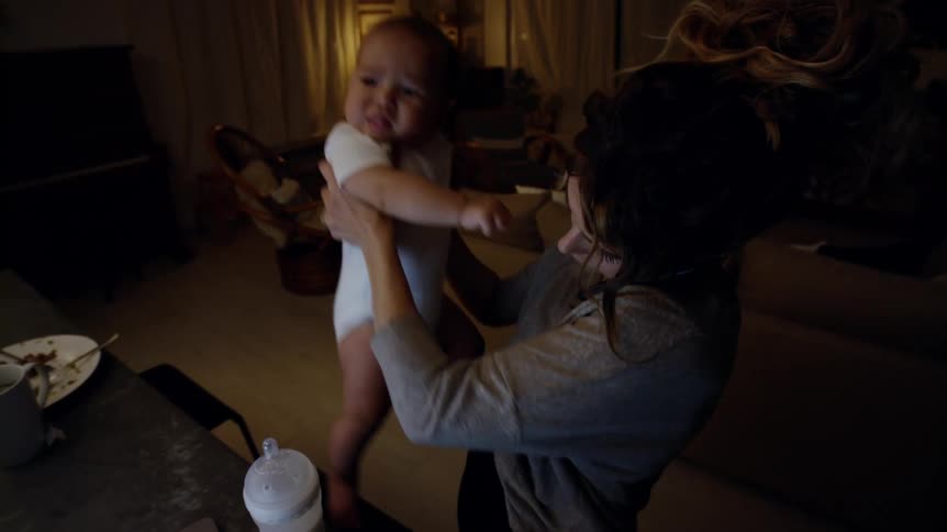 Mother with brown wavy hair and gray sweatshirt holds baby and talks on smartphone in living room at home at night