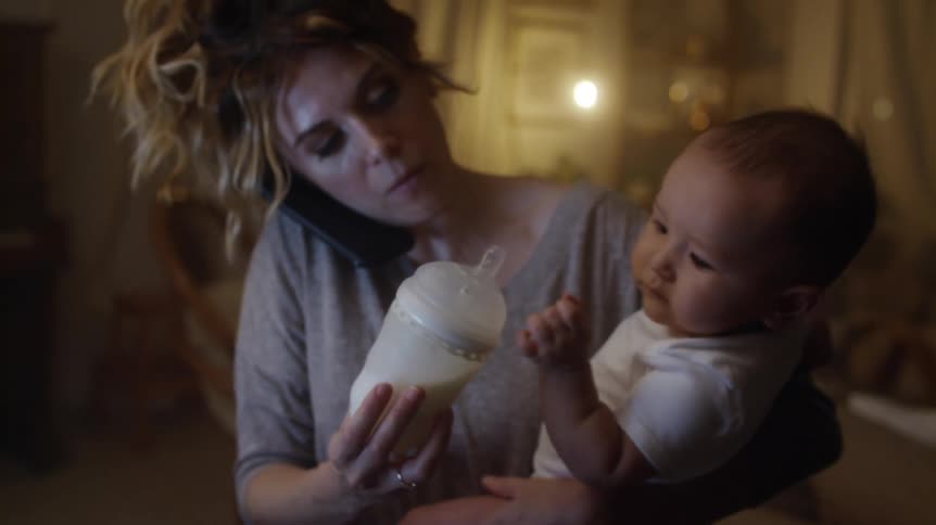 A frazzled looking woman tries to write notes while on the phone as she offers a bottle to the baby that she is holding