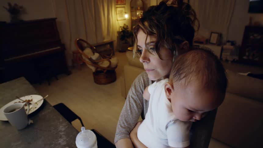 Mother patting her babys back while standing near baby bottle sitting on a table puts smartphone to her ear
