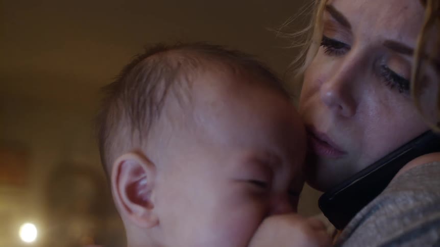 Mother with brown wavy hair and gray sweatshirt holds crying baby while talking on smartphone