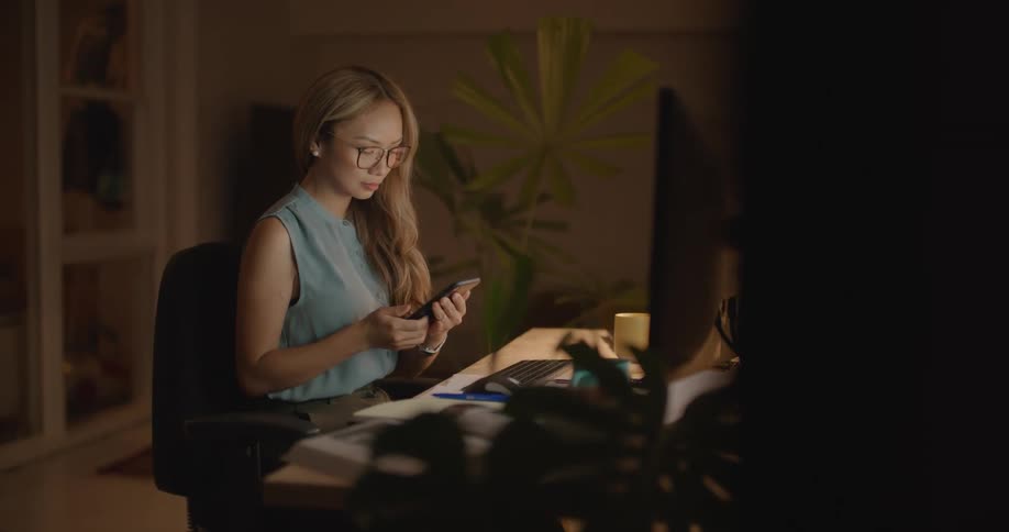 Woman with long wavy hair glasses and blue blouse receives call while working on computer in room