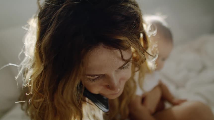 Mother with brown wavy hair sitting on bed holds baby while talking on smartphone
