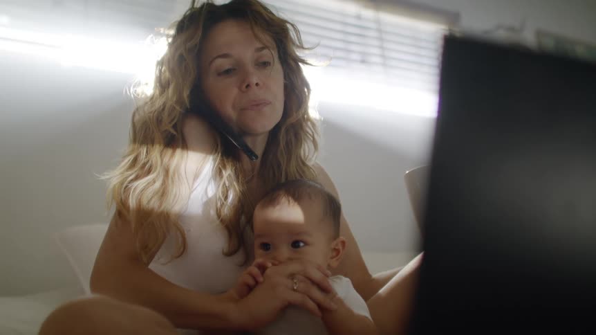 Mother sitting in the bed with her son in her lap talking to the phone while shes holding a cup and looking at the laptop screen