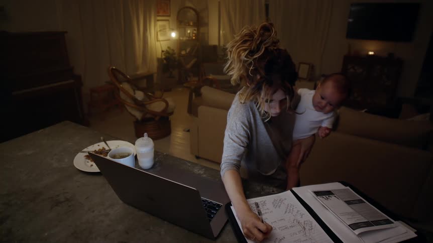 A harried looking mom standing at the kitchen counter holding baby while making notes working on a laptop and talking on the phone