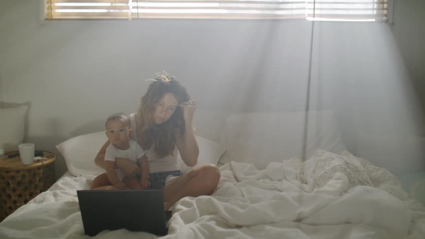 Mother with brown wavy hair white shirt and shorts sits on bed holding baby and making call on smartphone