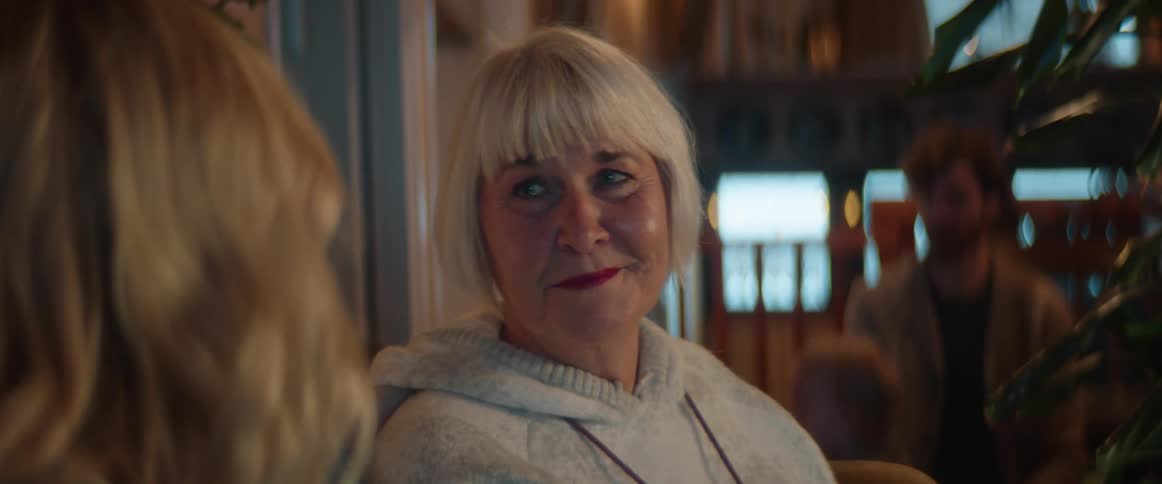 Woman with short blonde hair smiles at another woman while a man plays with a toddler behind her