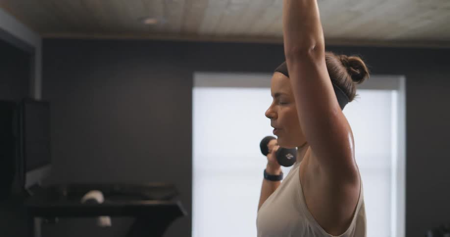 Woman with black headband wireless earphones and gray sport shirt lifts dumbbells in the gym
