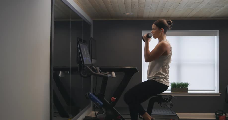 A young woman lifts weights while riding a stationary bike at home