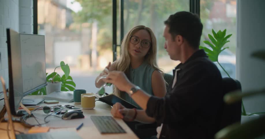 Man and woman sitting at the office desk in front of two computers analyzing a sunglasses and talking to each other