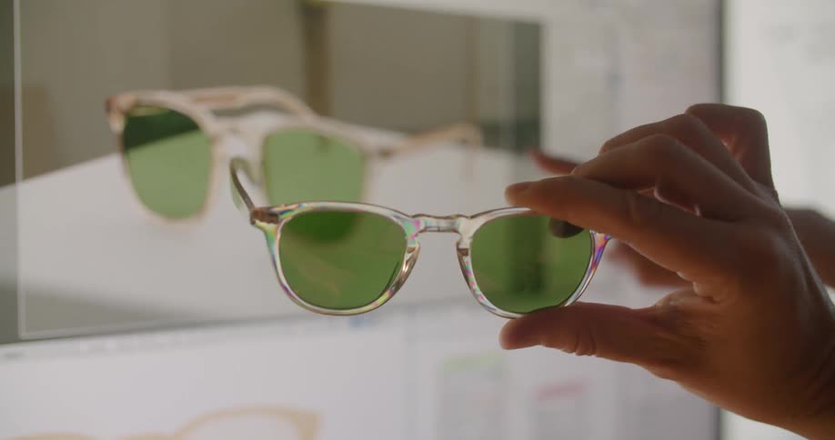 A person holds a pair of sunglasses up as another person points at a computer screen and compares them to the ones on the screen
