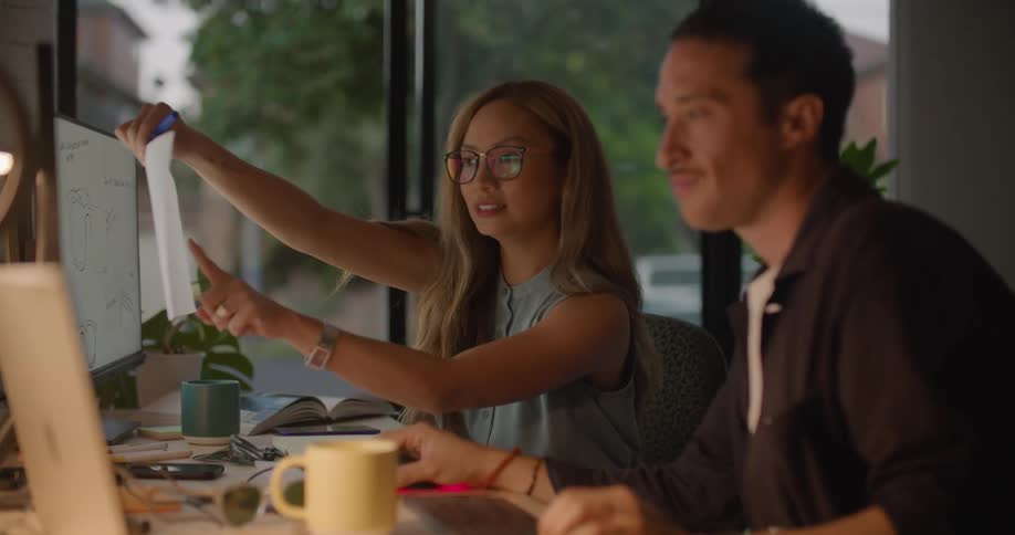 Woman holds a paper up in front of a monitor and smiles as a man sitting next to her looks at the paper and smiles too