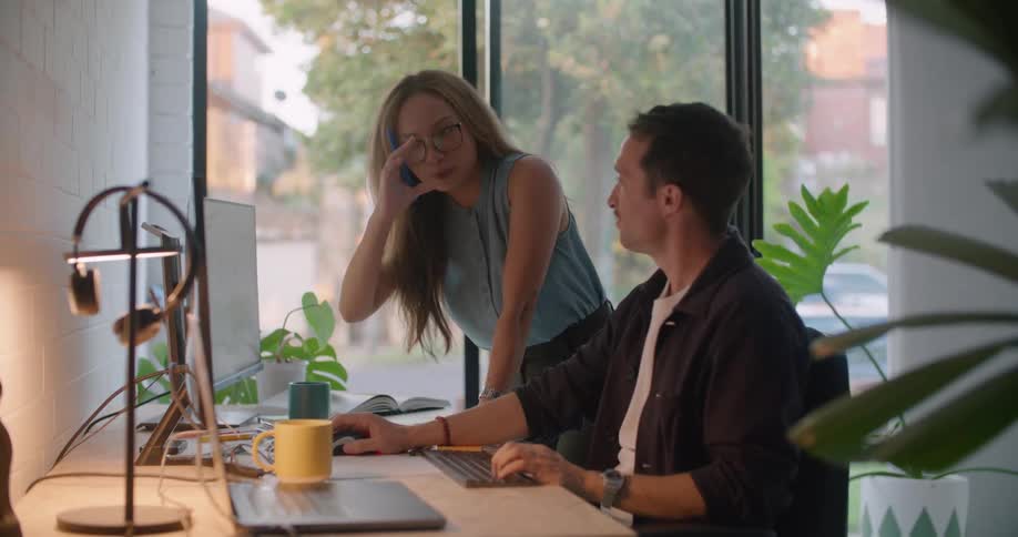 Man with white and black shirt sits at desk using computer and talking to woman with brown hair and blue blouse in the office
