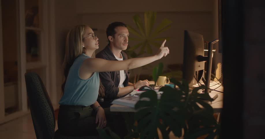 Man with white and black shirt sits at desk using computer and talking to woman with brown hair and blue blouse in the office