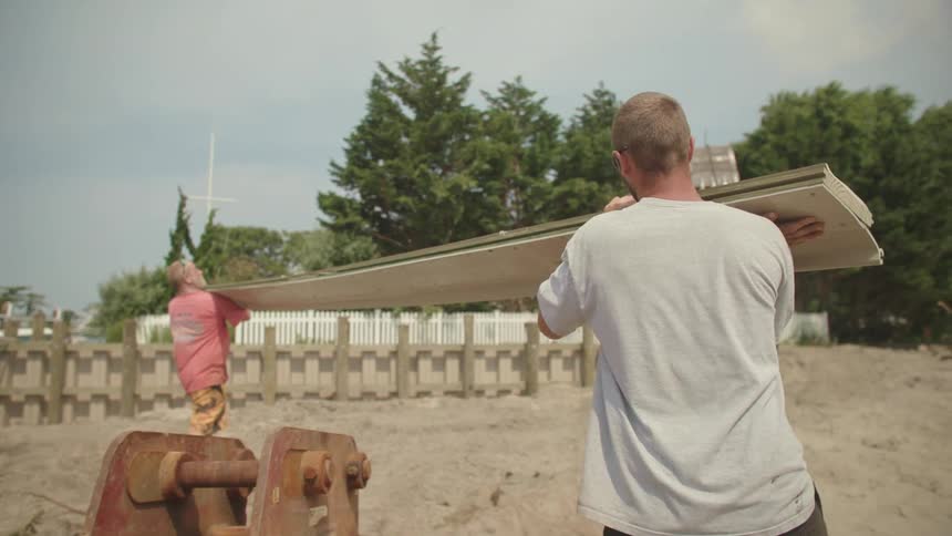Two white men carrying a pile of wooden boards in a houses construction site at a sunny day