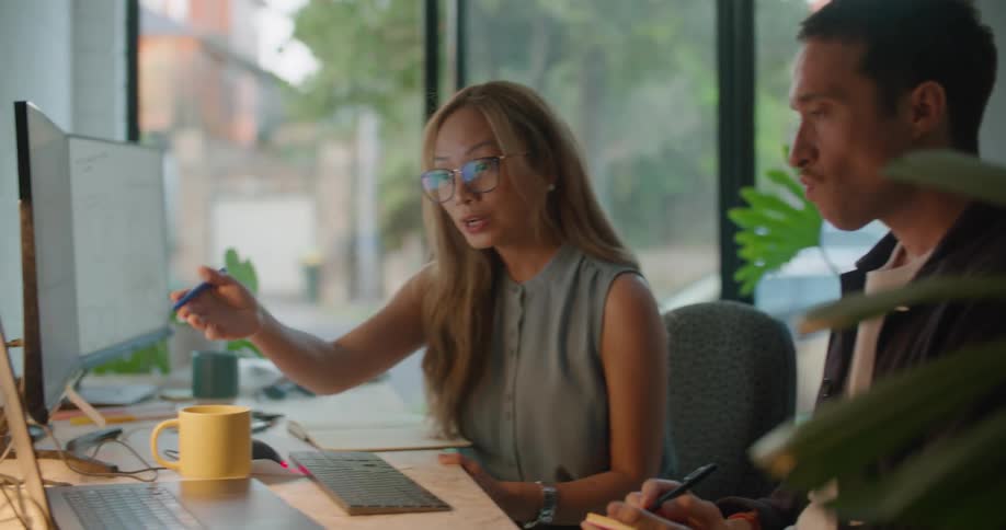Man and woman sitting at the office desk when she points at the laptop screen and talks to the man while he takes notes on his notepad