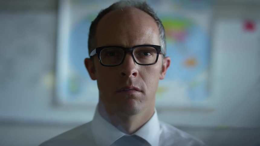 Teacher standing in front of the whiteboard and world map of a classroom wearing glasses and formal clothing
