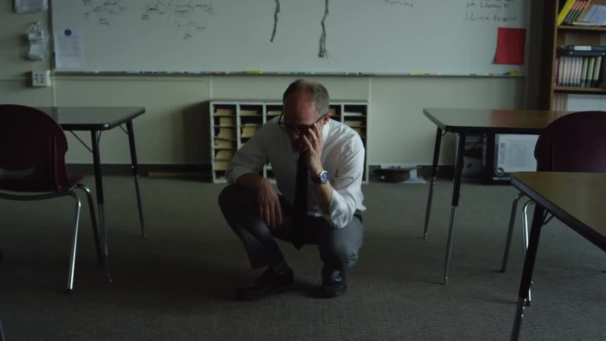 Man wearing a shirt and tie crouches on the floor in a classroom