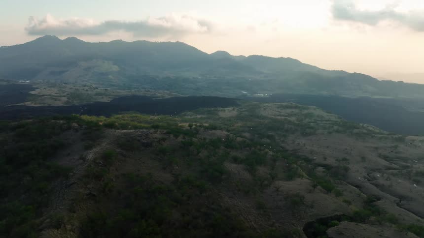 A flock of birds fly over a mountain and there are silhouettes of mountains in the distance
