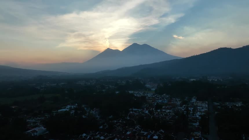 A neighborhood with silhouettes of Volcanos in the distance and the sun shining behind clouds in the sky