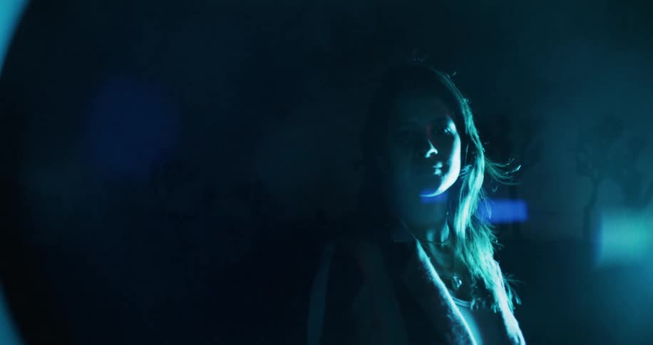 A woman looks at the camera while standing in front of a bright light at nighttime