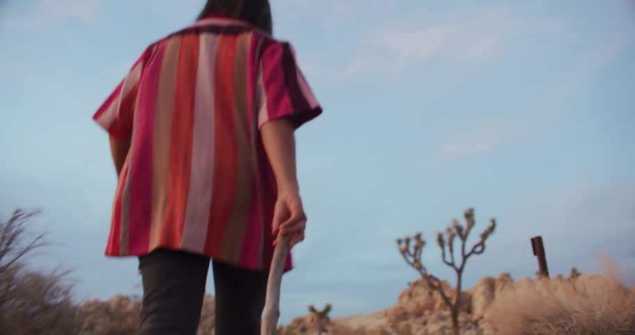 A woman walks through a desert while dragging a stick through the sand behind her