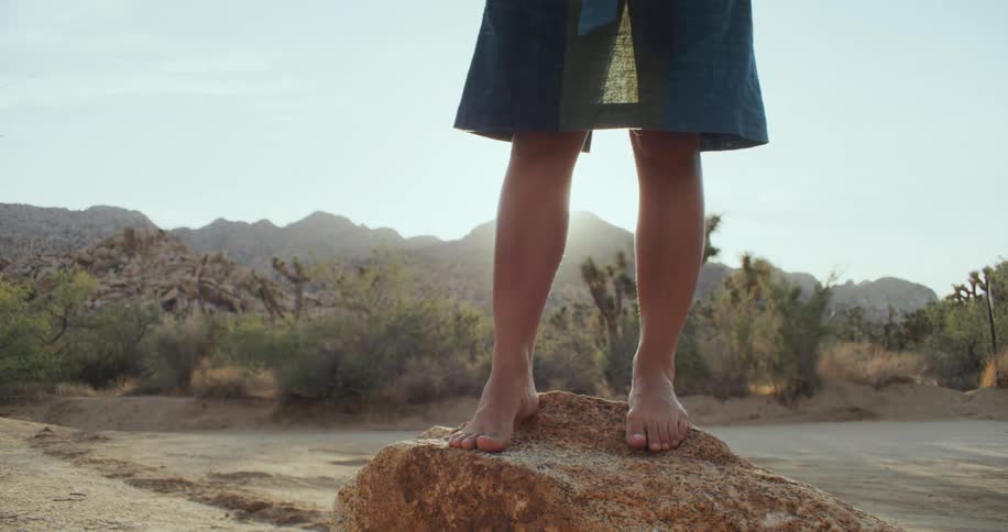 Person wearing skirt stands on boulder in sand near scrub land as sun back lights them from over mountain