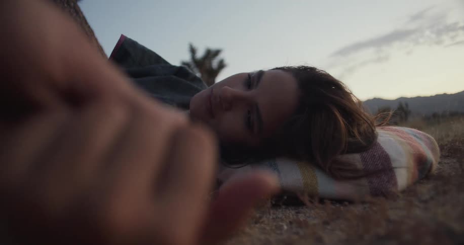 Woman with a sad looking face lying down in the ground of an outdoors field and having her hand touched by someone else in a romantic way
