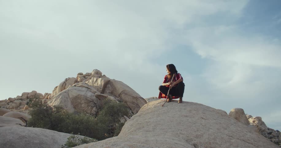 A woman crouches atop a large rock formation as she holds a stick in her hands