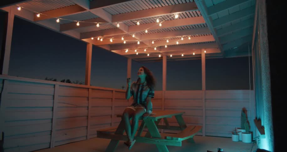 Woman sitting on the table at the houses balcony turning the hanging lights above her on and off while watching in awe