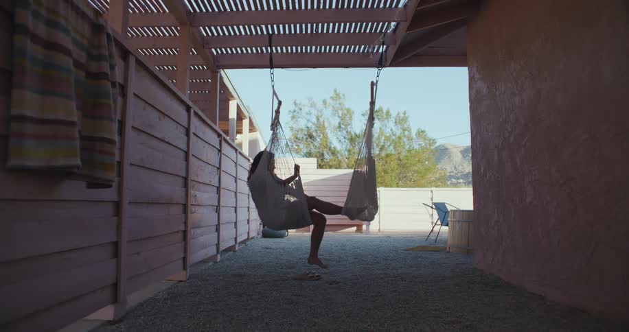 A woman in a hanging hammock chair reaches out for the foot rest and props her feet in it