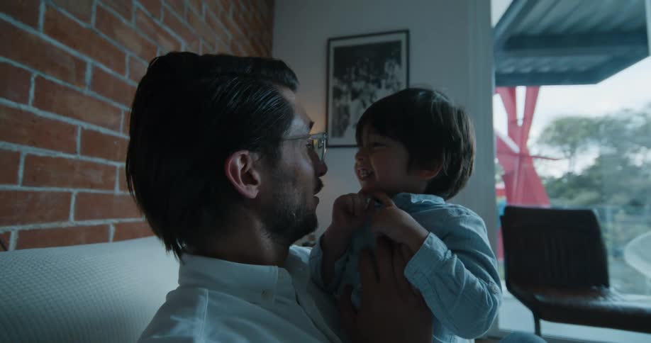 Father sitting in the couch in his living room holding his son and talking and laughing with him