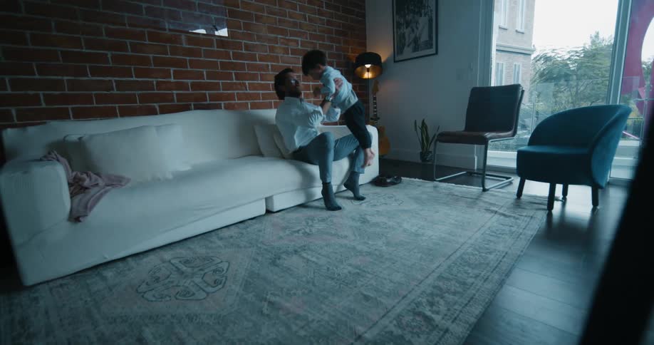 Toddlers run to their father who is sitting on a white sofa and he lifts them into the air