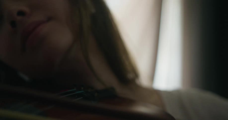 White woman playing violin with closed eyes next to a big window