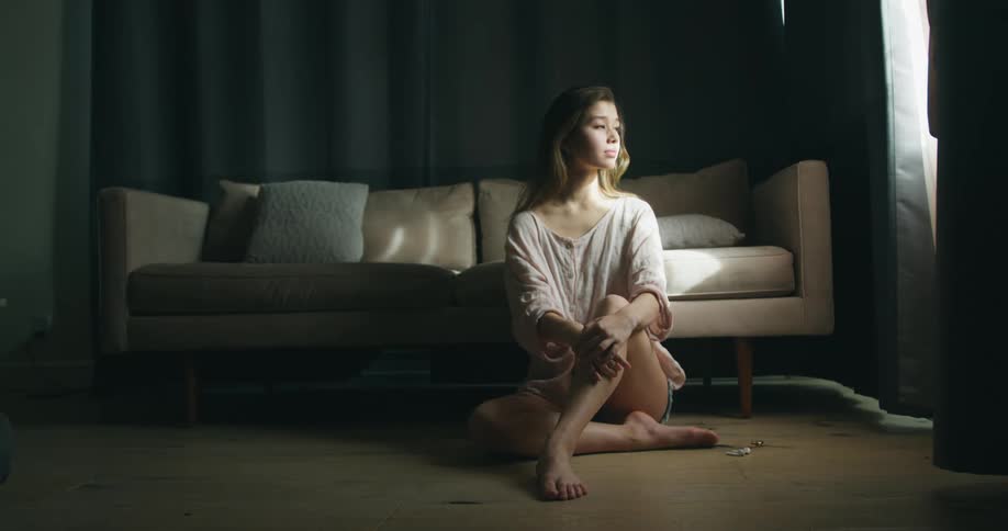 Young woman sitting on the floor alone in front of a couch and looking through the window