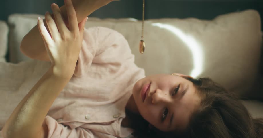 A teenage girl lies in bed and holds a locket in front of her and stares at it as it spins