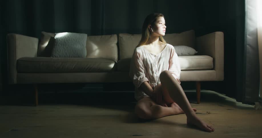 Young woman wearing a wireless earphone sitting in the floor in front of a couch and looking through the window on her side