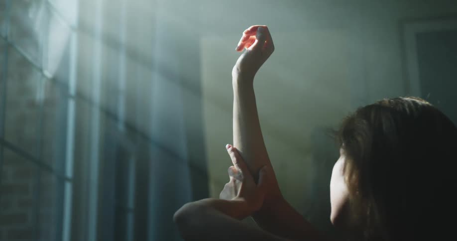 A woman stands in front of an open window and raises her arm while bathed in sunlight