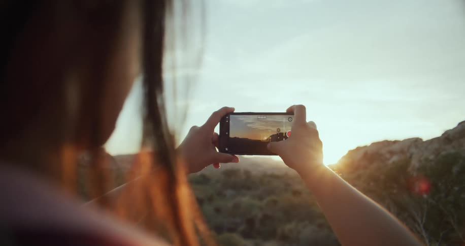 Woman standing near a forest taking pictures of the sunset ahead with her smartphone