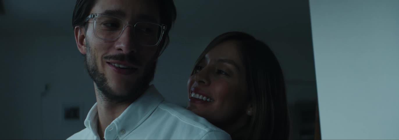 Woman hugging her husband from behind while they talk and smile inside the house next to a glass window