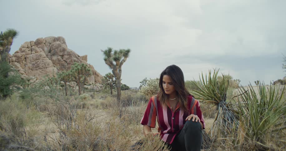 Woman half kneels in scrubby desert examining plants and looking around