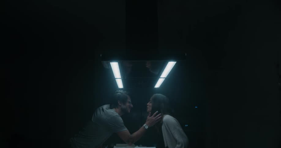 A couple lean in to kiss in a dark barren room
