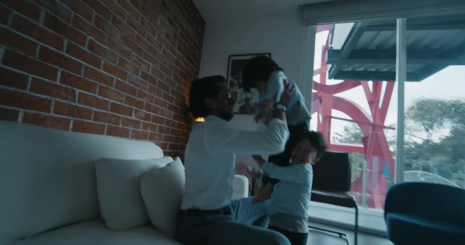 Father sitting on the couch playing with his two sons that come running towards him lifting one of the boys up in the air