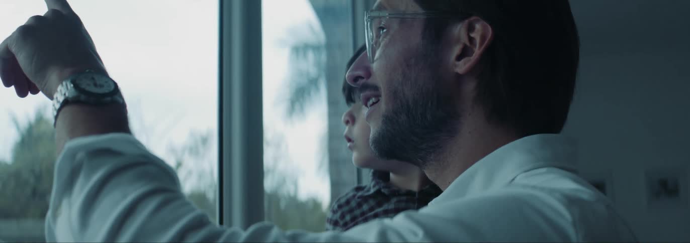 A father holds his young son in his arms while talking to him as they look out a large window