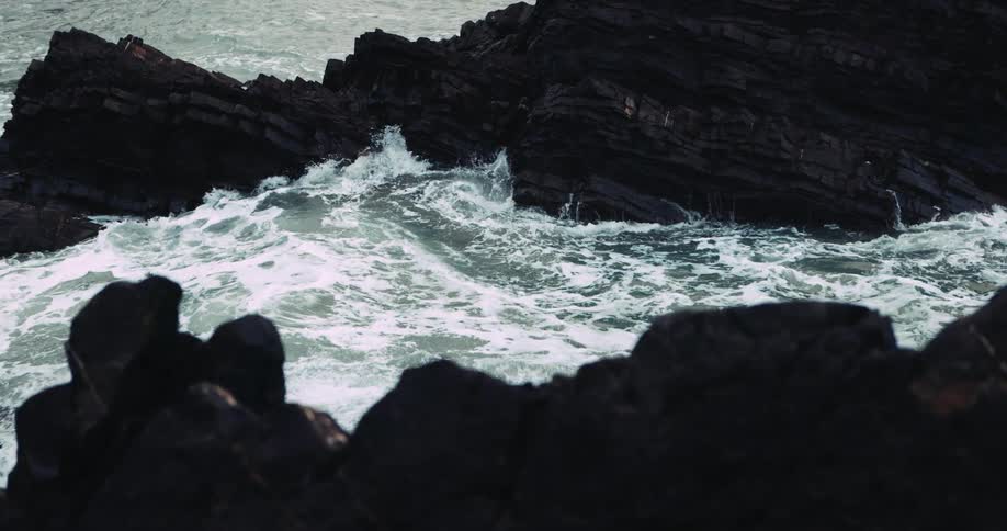 A tall wave rises up in the ocean and crashes onto rocks on the coast