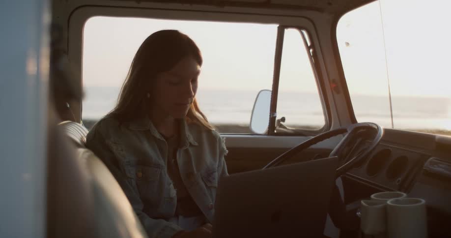 A woman sitting in the drivers seat of a truck typing on a laptop parked near the ocean
