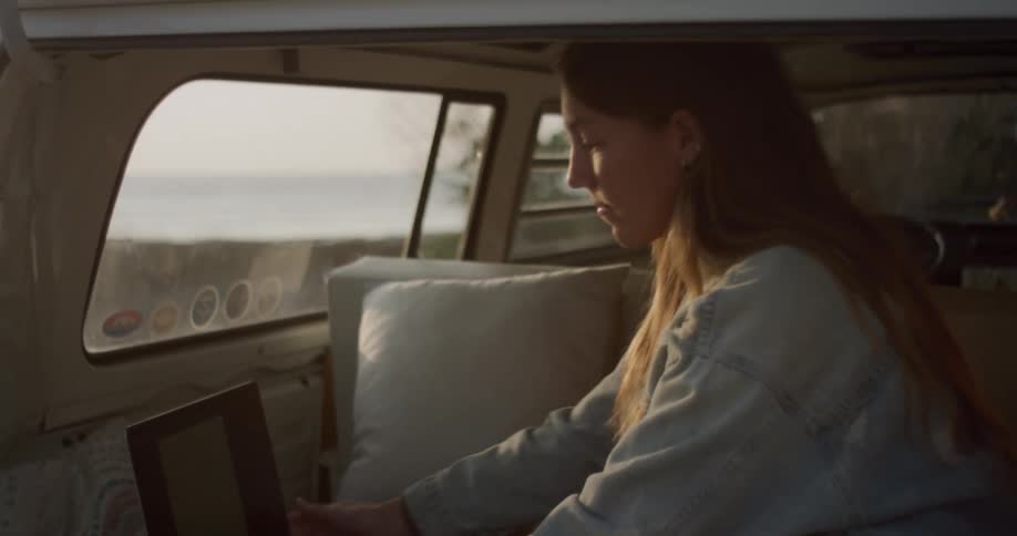 Woman types on a laptop while sitting in the back of a van that is parked near the ocean