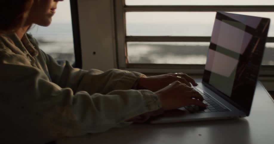 A young woman types on a laptop while sitting at a table in a recreational vehicle parked on the coast during the day