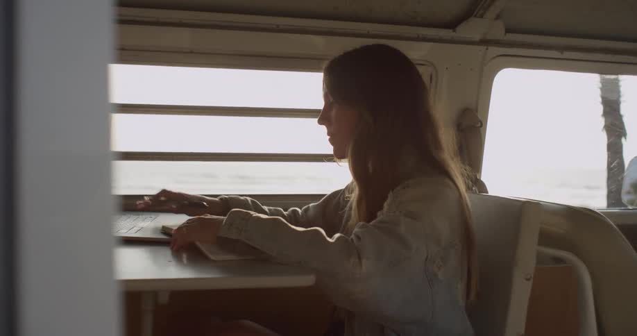 A woman sitting in a van near the ocean working on a laptop making notes