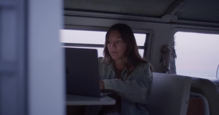 Woman sits in back of van typing on laptop with side window showing view of ocean