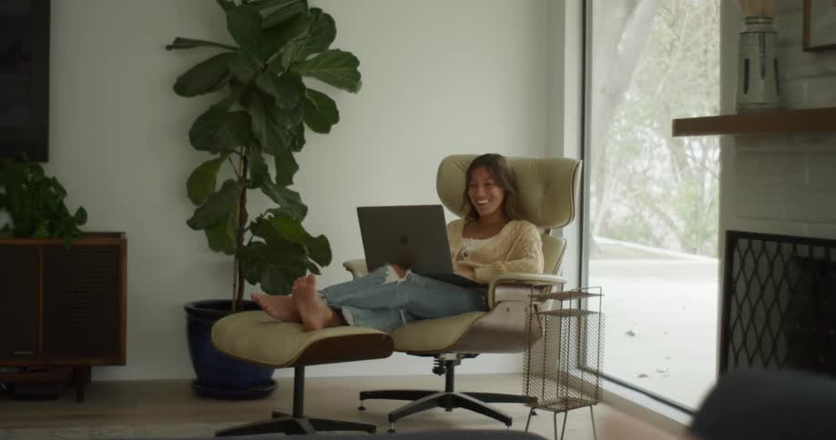 Young barefoot woman laughing while sitting with a laptop on a chair in the living room